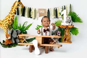 Safari themed first birthday setup with stuffed animals, decorative banners, and tropical foliage on a white backdrop.