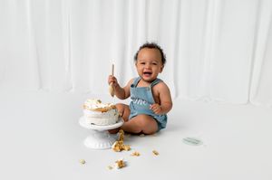 Happy baby in blue romper sits on white floor with smashed birthday cake during first birthday cake smash photo session.