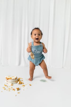 Baby in blue outfit sits on white backdrop with cake crumbs scattered around during first birthday cake smash photoshoot.