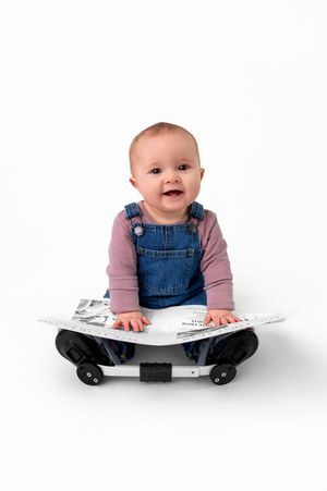 Baby in denim overalls and pink shirt sits happily on a skateboard against white background.