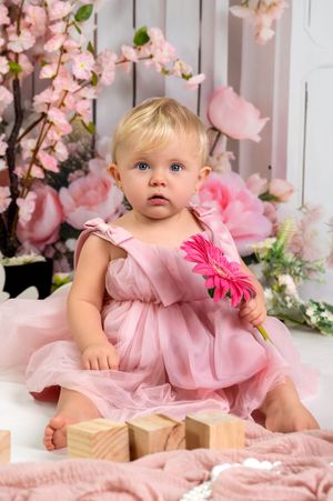 Sweet baby in pink dress poses among cherry blossoms and floral decorations for spring portrait session.