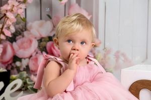 A small child in a pink dress sits among floral decorations and soft pink backdrops for a portrait session.