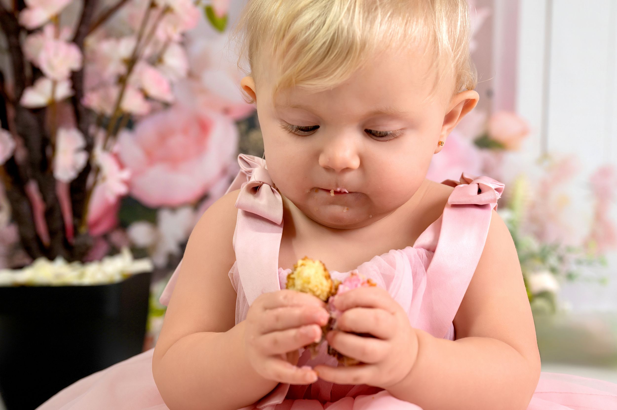 A toddler in a pink dress sits among delicate cherry blossoms and spring flowers in a dreamy portrait setting.