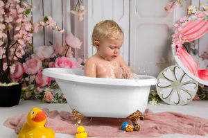 A pink and white themed baby photography setup with a vintage bathtub, rubber ducks, and floral decorations.