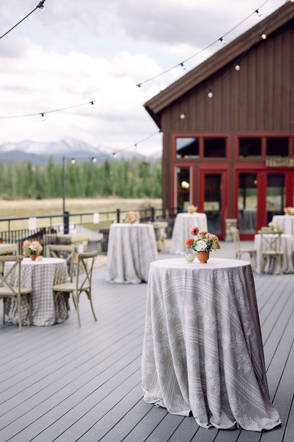 Outdoor wedding reception setup with round tables covered in gray linens on a wooden deck with mountain views.