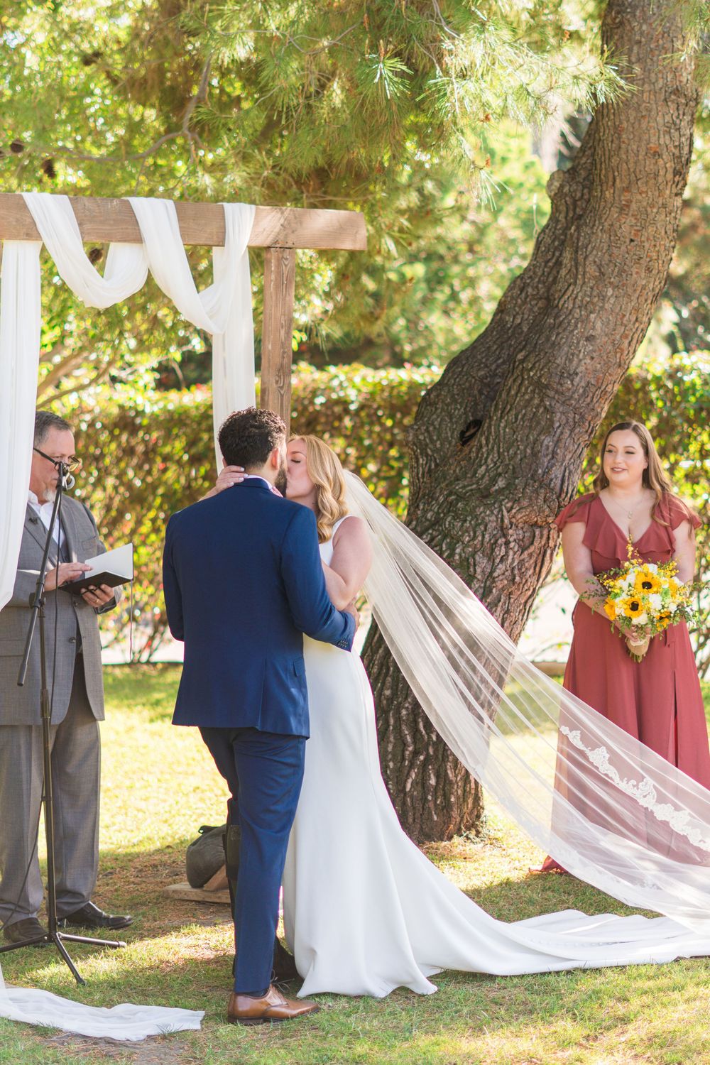 A wedding ceremony takes place outdoors under tall trees with flowing white drapery and guests in formal attire.