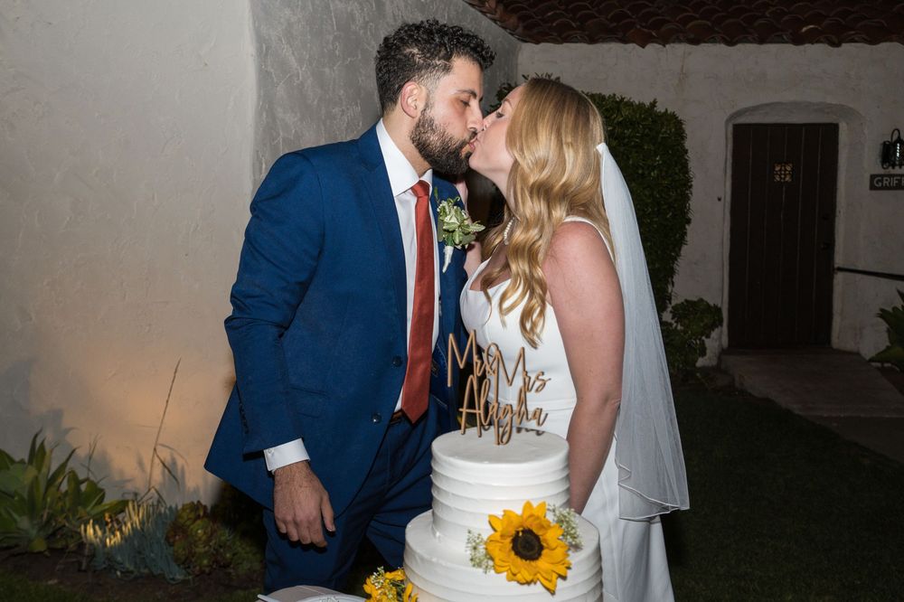 A couple in wedding attire share a romantic moment while holding a sunflower bouquet against a stucco wall.