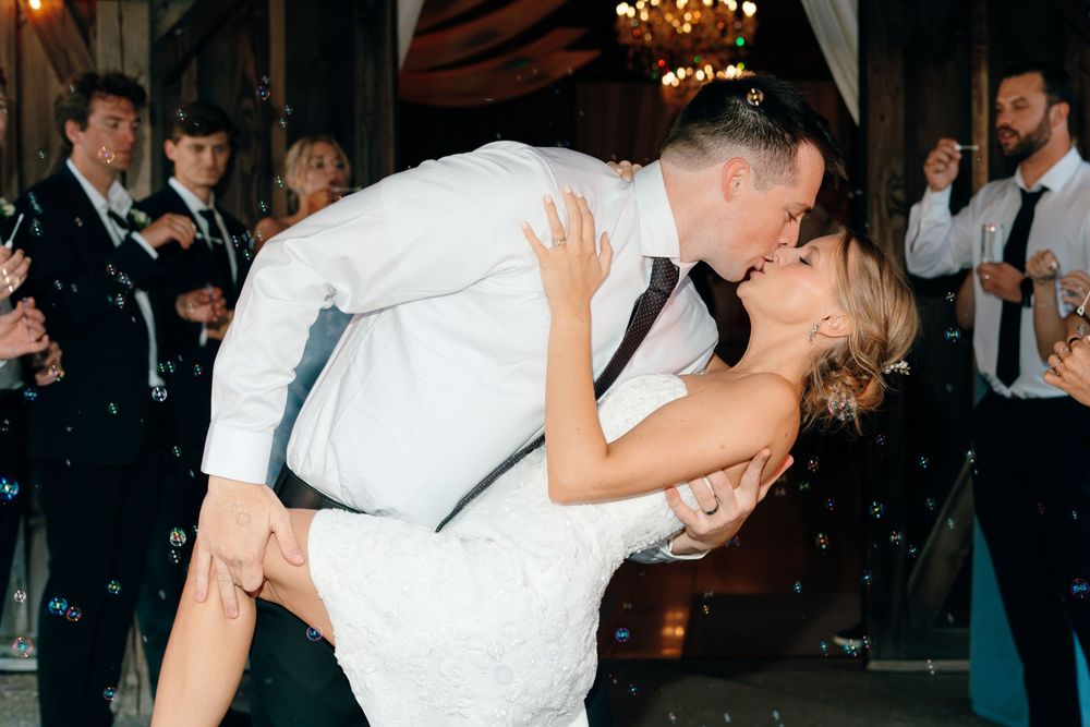 Groom dipping bride for romantic kiss on dance floor surrounded by wedding guests.