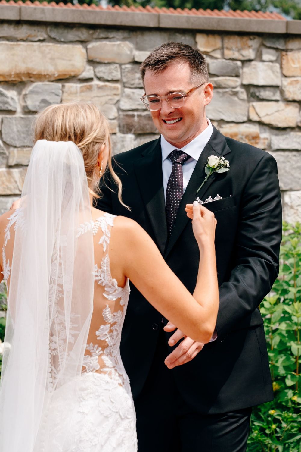 Groom smiling joyfully at bride during first look, wearing black suit with white boutonniere, stone wall backdrop.