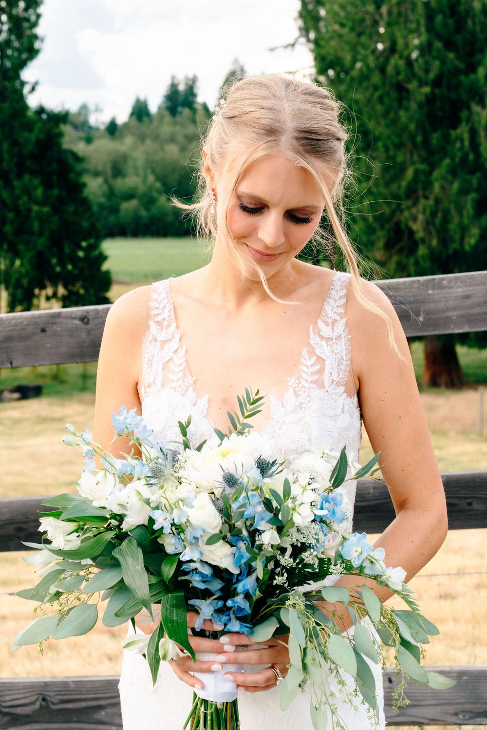 Blonde bride in lace wedding gown admiring blue and white floral bouquet outdoors.
