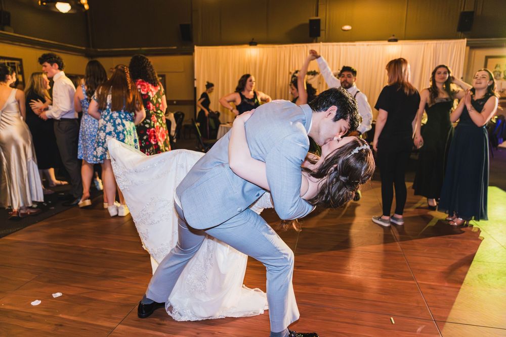 Wedding guests dancing and celebrating on a wooden dance floor at an indoor reception venue with warm lighting.