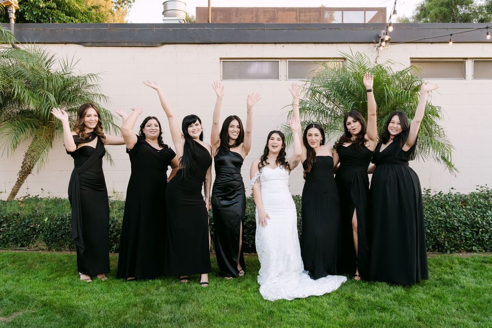 Wedding party wearing black and white attire poses together on grass near palm trees and building exterior.