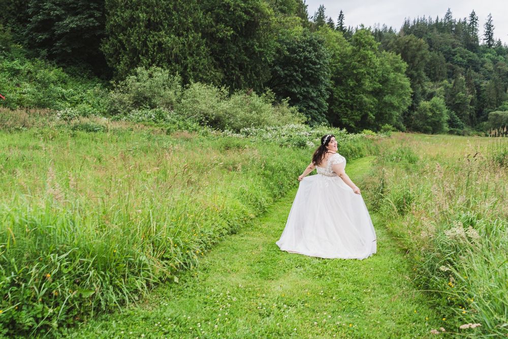 A person in a flowing white wedding dress poses gracefully in a lush green meadow surrounded by forest scenery.