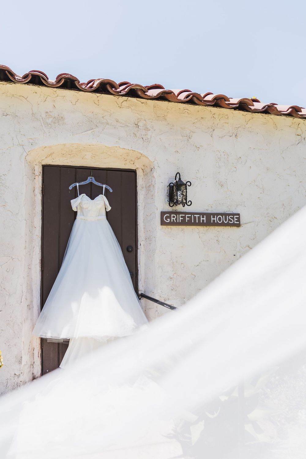 A white wedding dress hangs outside the Griffith House against a white stucco wall with Spanish tile roof.