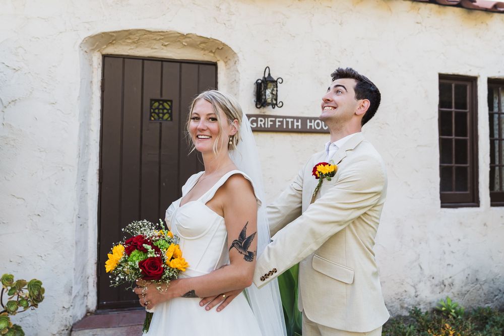 Wedding couple laughs together against white stucco wall of Spanish-style building.