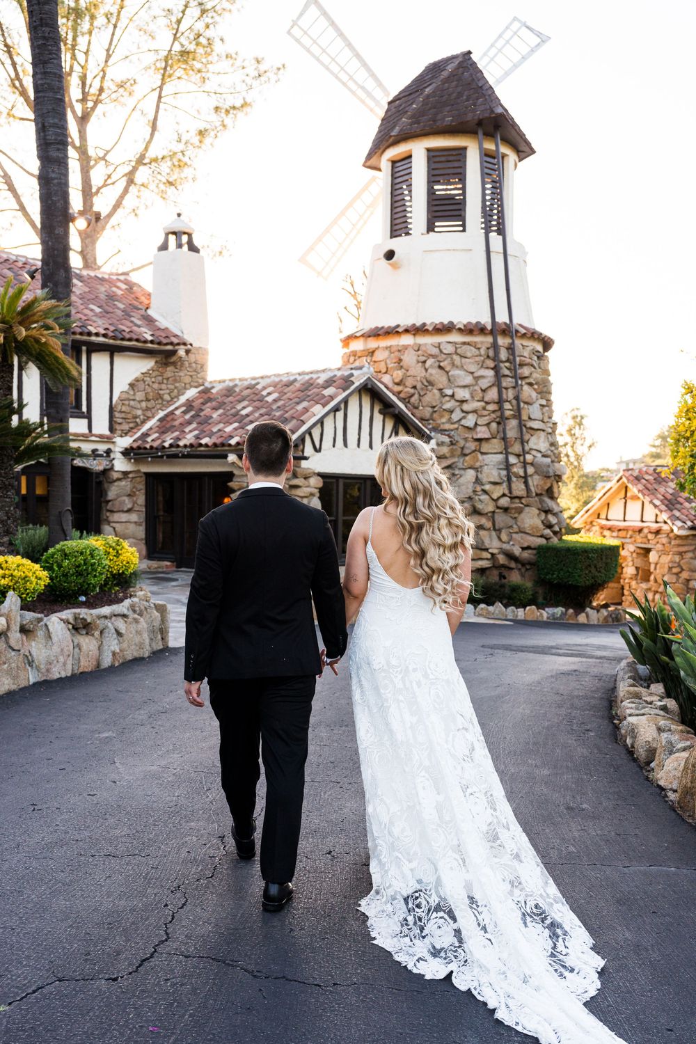 A couple in wedding attire walk down a stone-lined path with turrets and towers visible in the background.