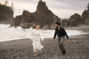A couple in formal attire walks along a dramatic rocky beach coastline with ocean waves and cliffs in the background.