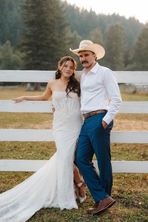 Bride in lace gown and groom in blue pants with cowboy hat pose by white fence at rustic outdoor wedding.