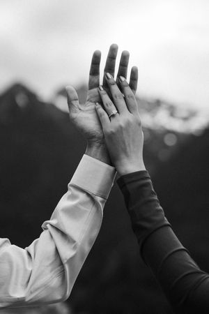 Black and white photo of two hands raised together against mountain backdrop, one wearing an engagement ring.