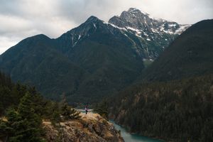 Person standing on rocky overlook with turquoise lake and snow-capped mountain peaks surrounded by dense forest.