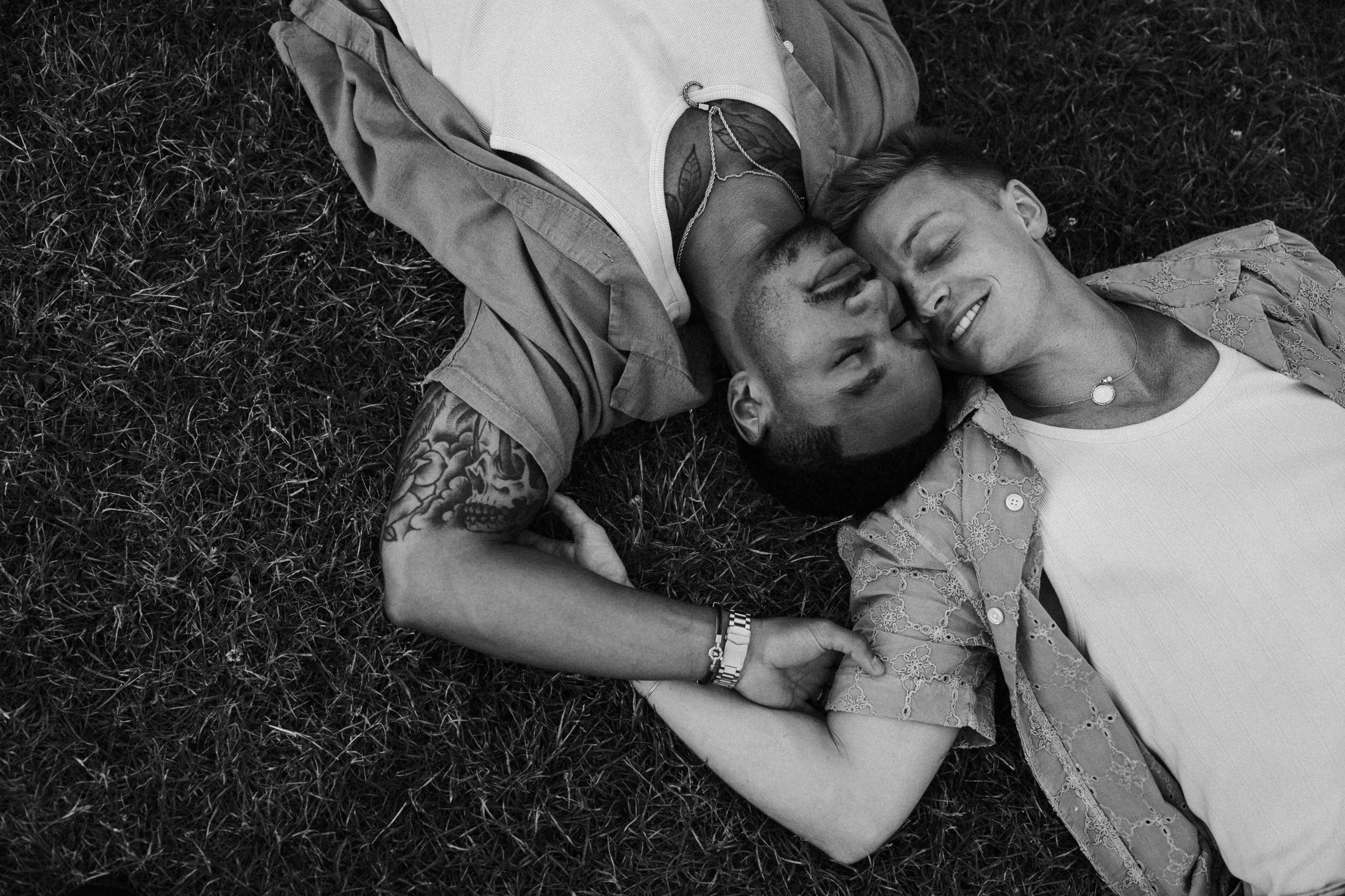Black and white photo of a couple lying on grass, smiling and holding hands in a romantic outdoor setting.
