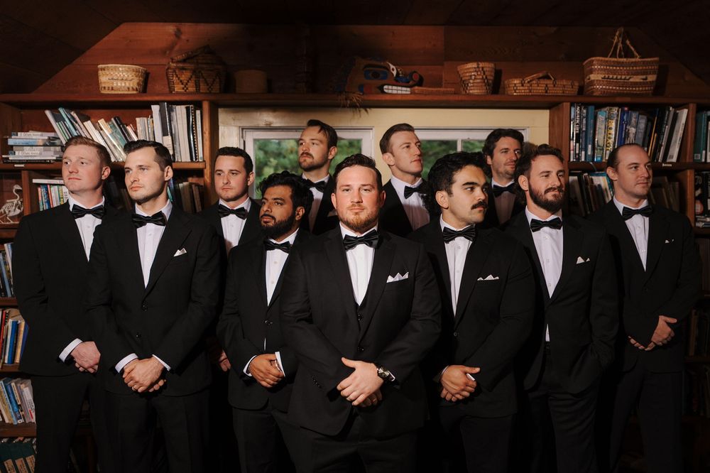 Group of groomsmen in black tuxedos, bow ties and white pocket squares posing together in a cozy library room with wooden bookshelves.