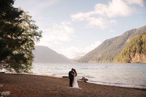 Bride and groom embrace on a pebbly lakeshore with mountains, calm water, and a large tree framing their elopement ceremony.