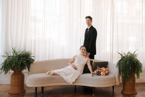 Bride reclines on a curved ivory sofa while groom stands behind, surrounded by ferns in a bright, airy wedding portrait.