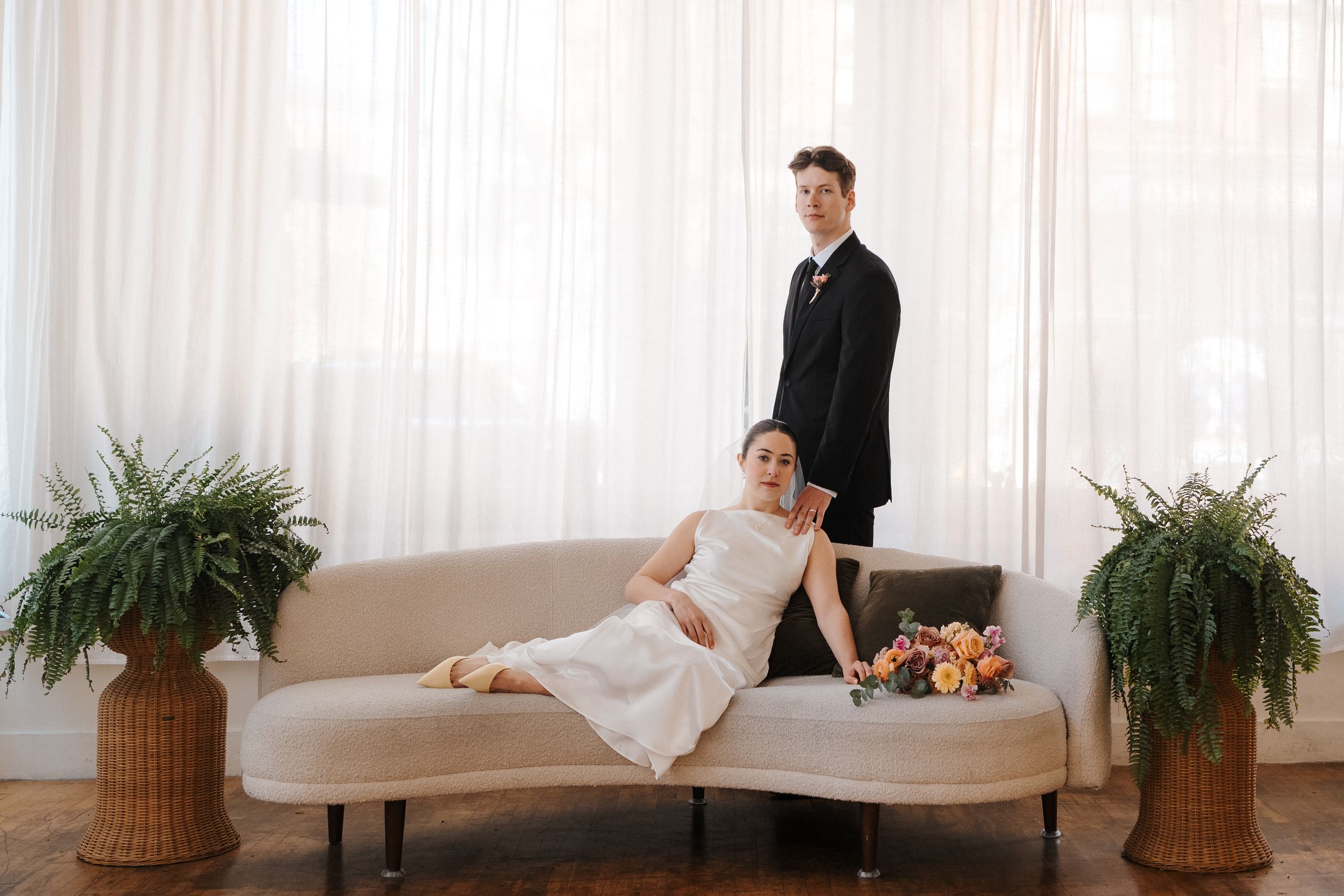 Bride reclines on a curved ivory sofa while groom stands behind, surrounded by ferns in a bright, airy wedding portrait.