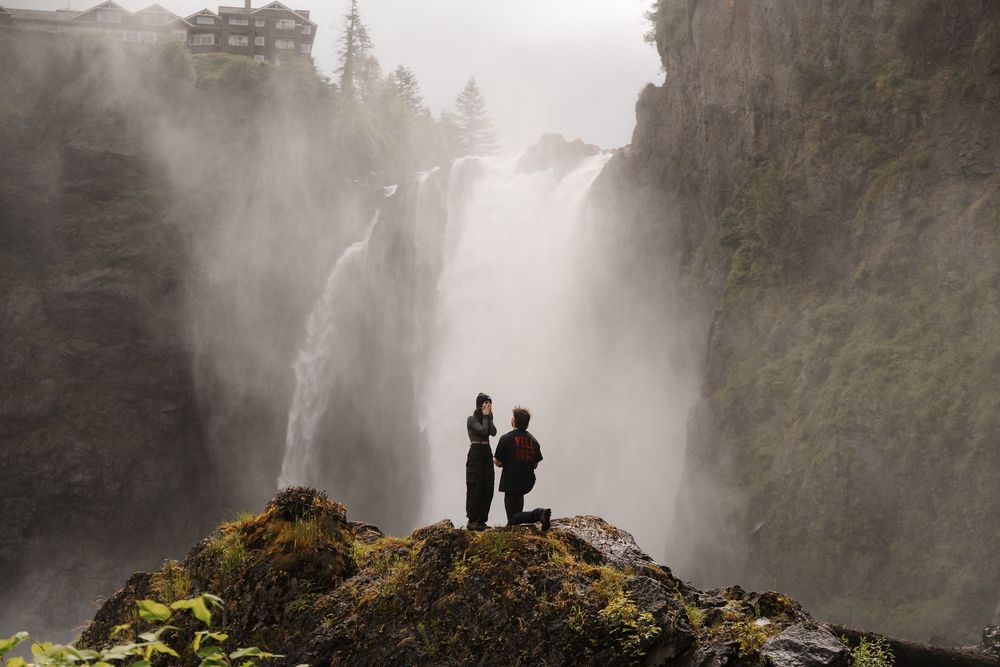 Couple stands on misty rock ledge before a dramatic waterfall during a romantic outdoor marriage proposal.
