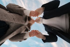 Two people wearing coats stand face to face and hold hands together against a blue sky background.