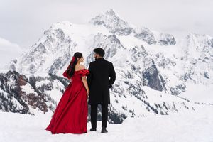 A couple in winter attire poses together against a dramatic snowy mountain landscape backdrop.