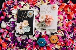 His and Hers wedding vow books with floral decorations and a ring box displayed on colorful rose petals.