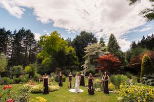 Bride and bridesmaids in dark gowns stand in a lush garden wedding ceremony surrounded by colorful trees and flowers.