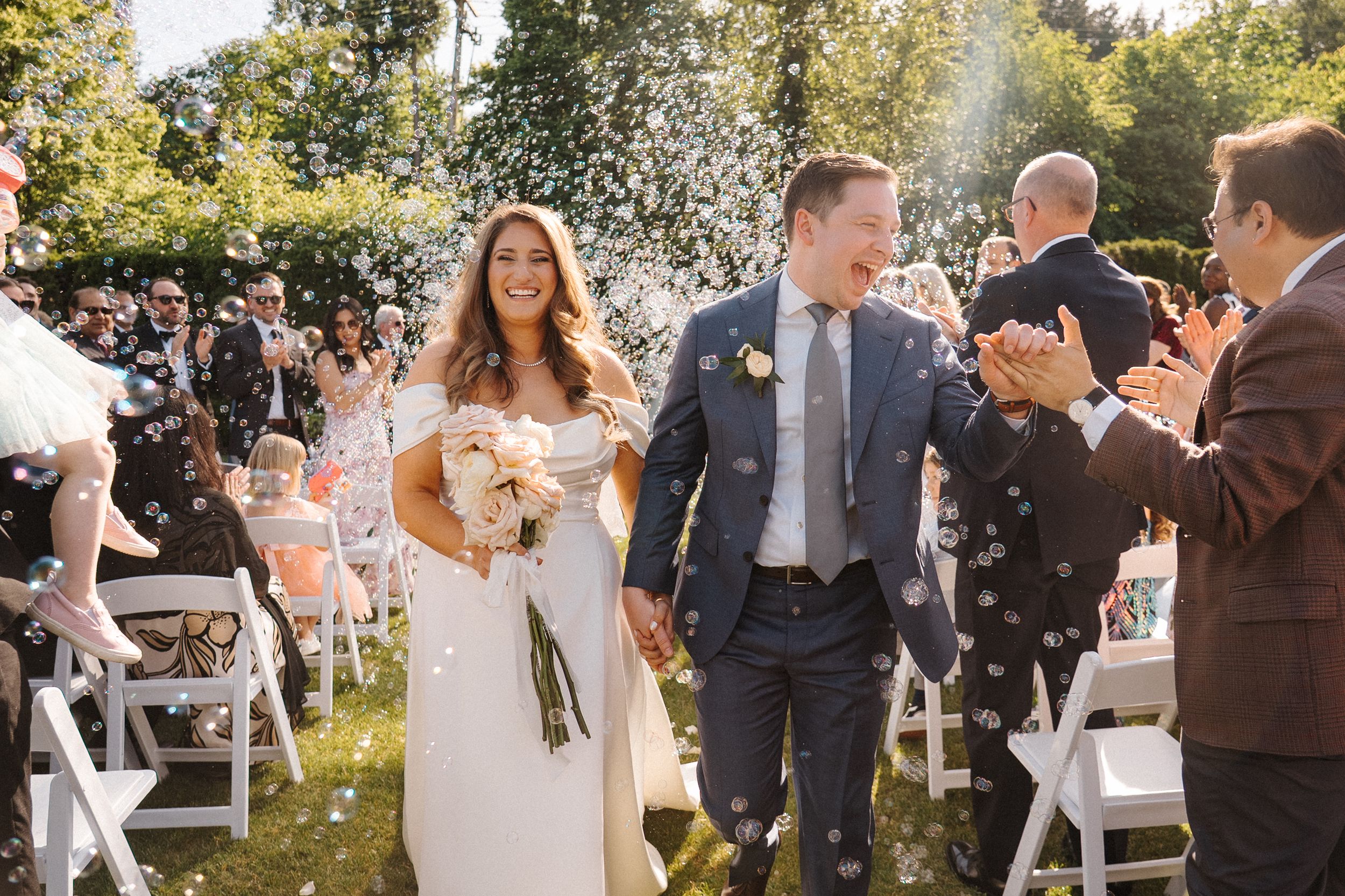 Wedding guests gather outdoors on a sunny day in a garden setting for a celebratory event.