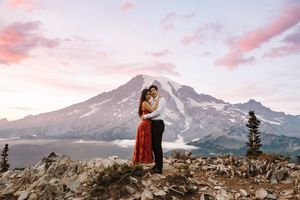 Two people embrace on a mountain peak at sunset with snow-capped peaks and dramatic pink clouds in the background.