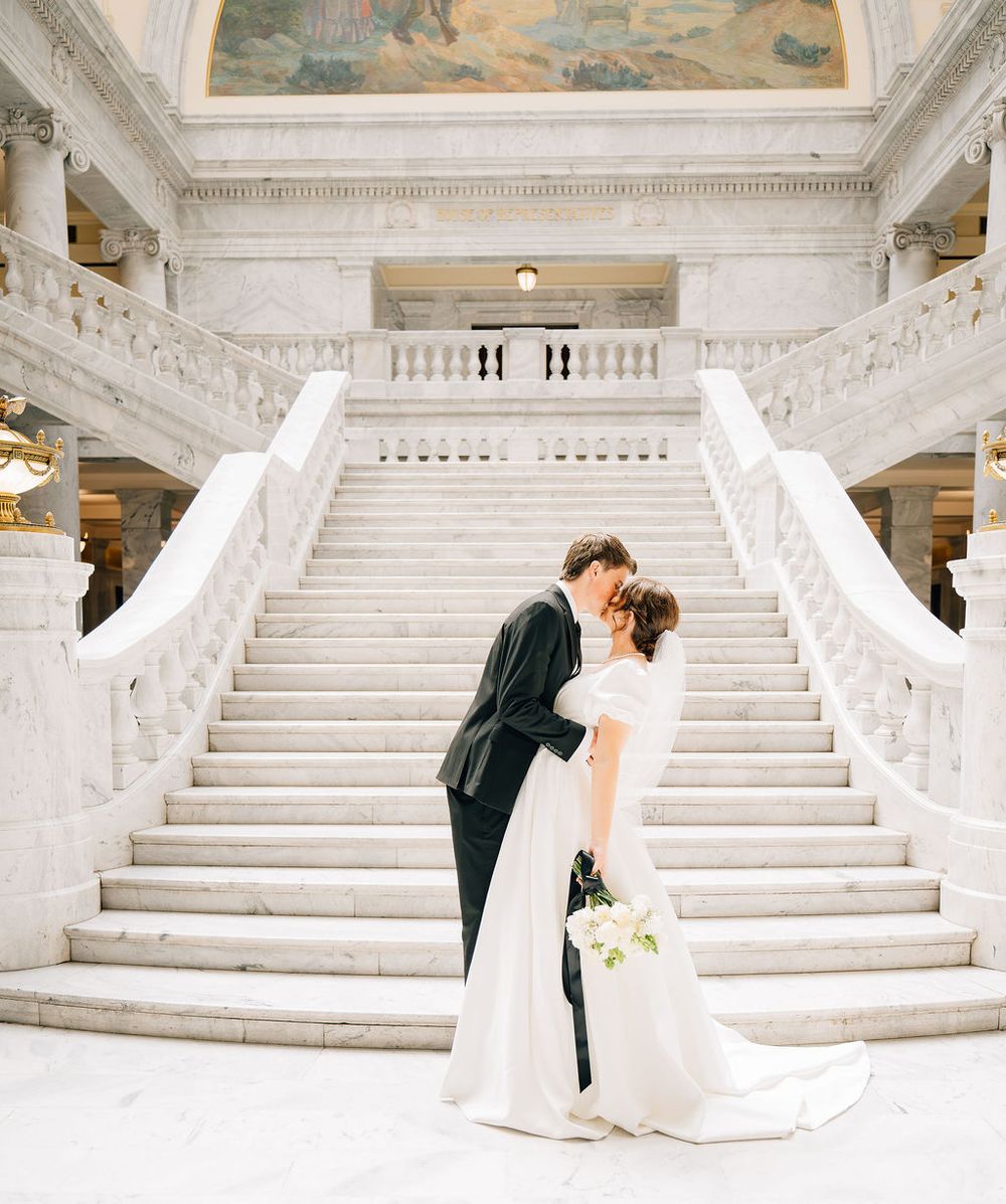 Bride and groom share a kiss at the base of grand marble staircase in elegant wedding venue.