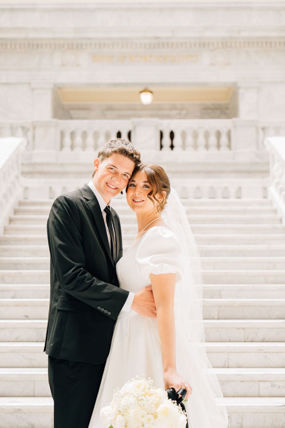 Smiling bride and groom embrace on marble steps at elegant wedding venue.