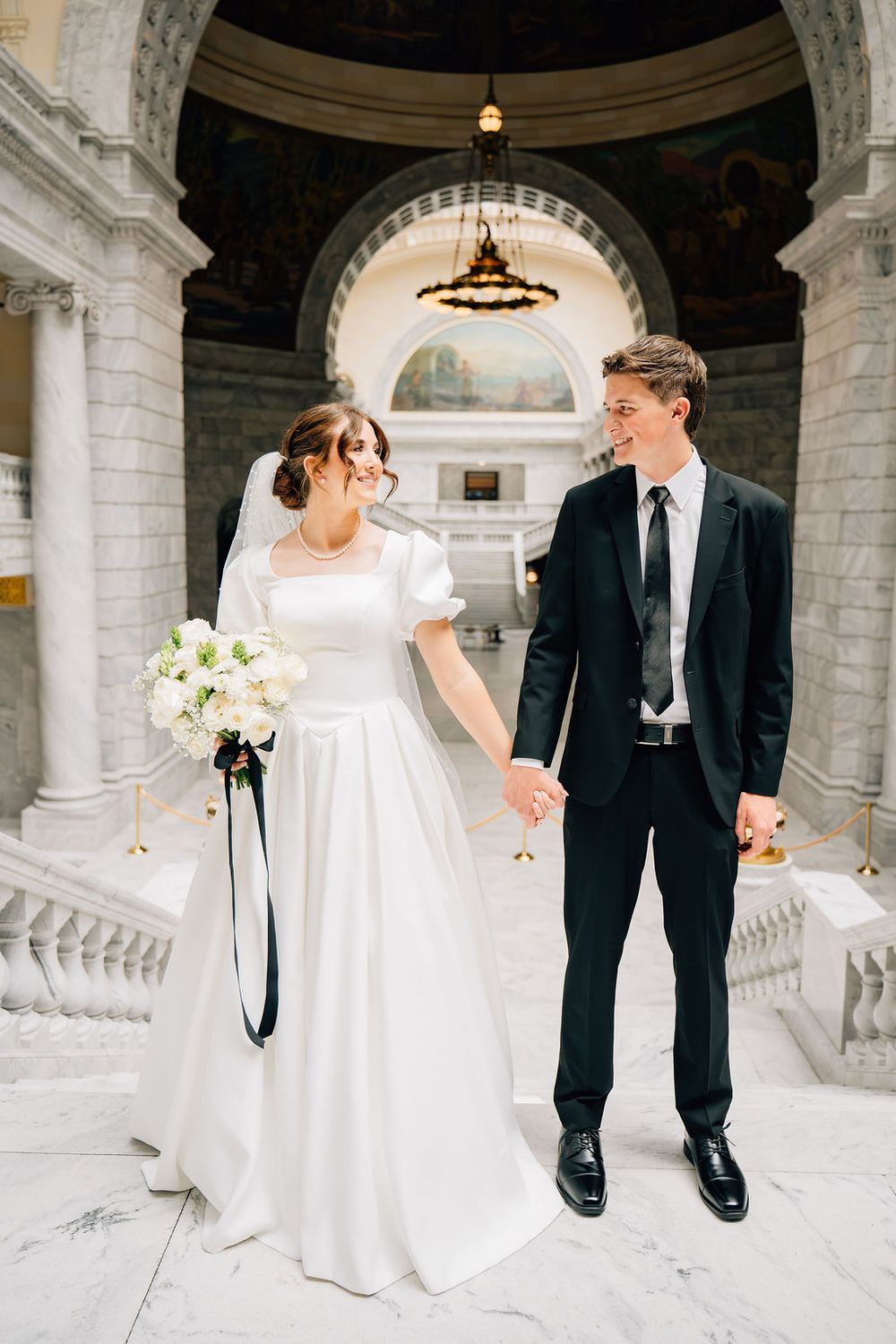 Bride and groom holding hands smiling in elegant marble rotunda wedding venue.