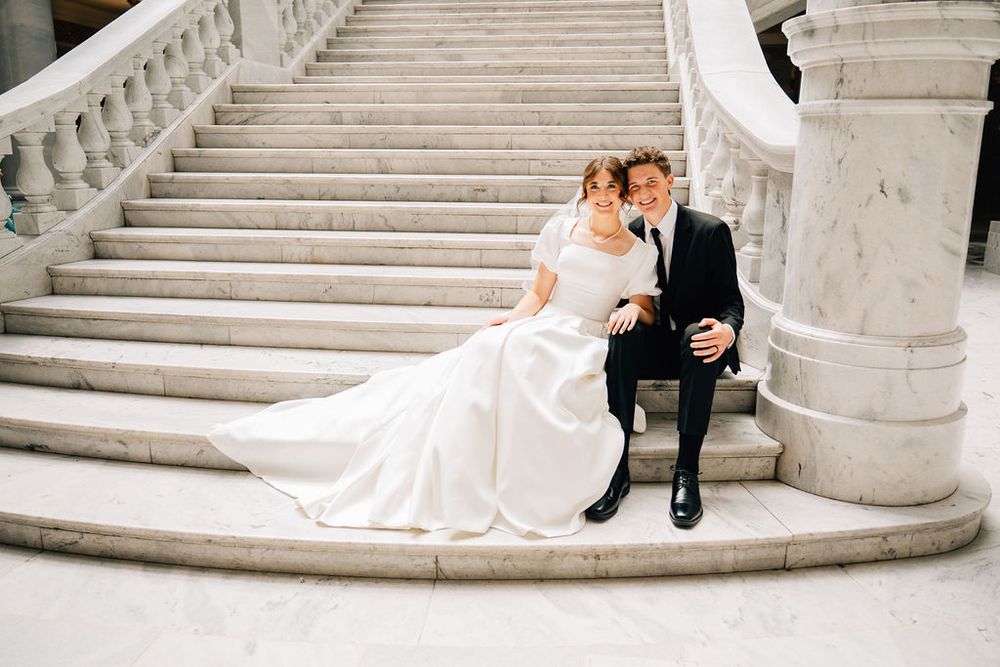 Bride and groom sitting together on elegant marble staircase, bride in flowing white gown, groom in classic black suit.