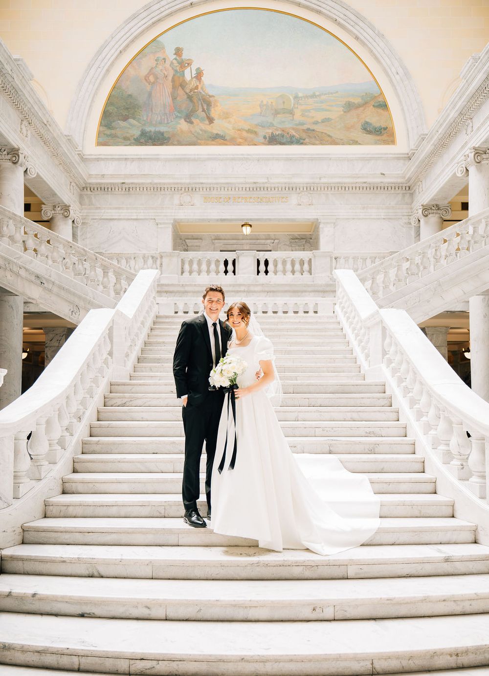 Bride and groom pose on sweeping marble staircase beneath ornate mural.