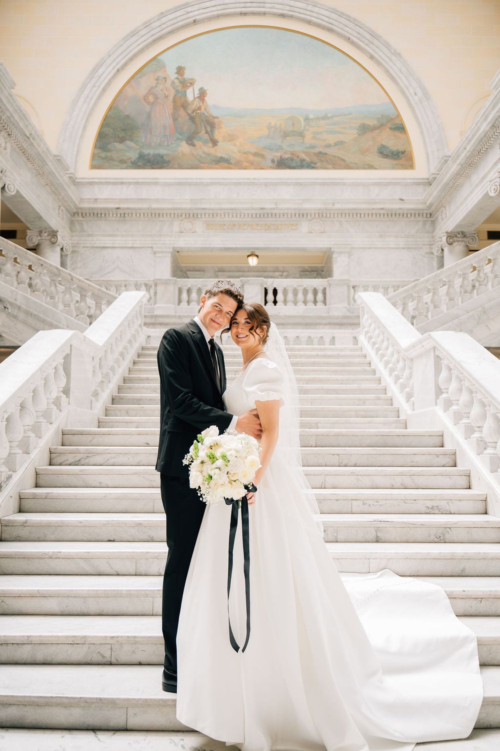 Bride and groom pose on grand marble staircase with classical mural backdrop holding white bouquet.