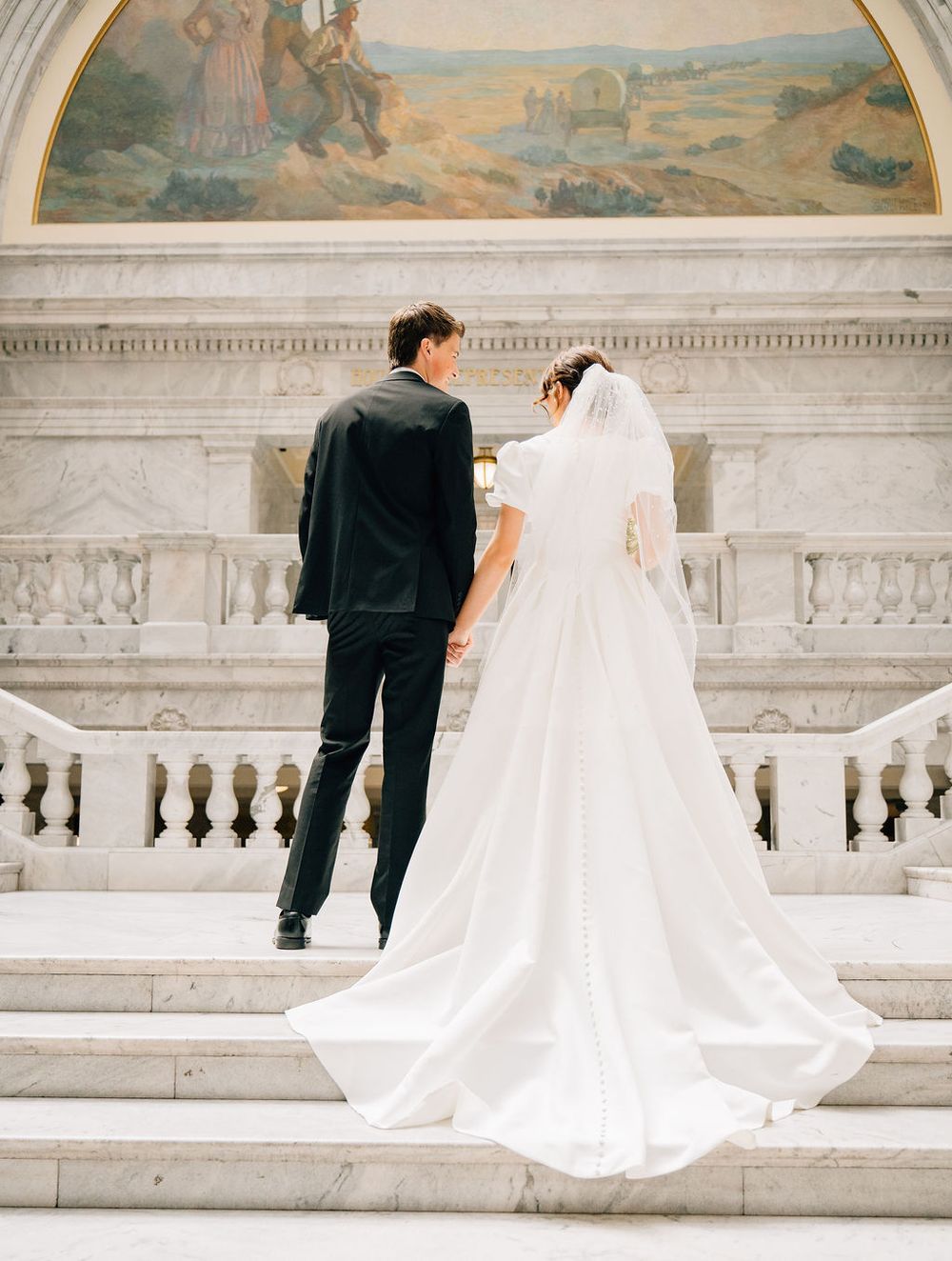 Bride and groom holding hands ascending marble stairs in elegant wedding attire at a grand classical building.