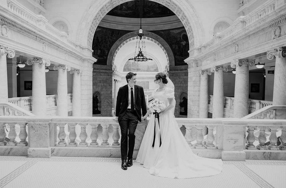Black and white wedding photo of bride and groom walking hand in hand in an elegant classical building.