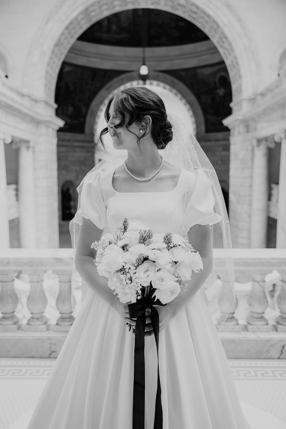 Black and white photo of a bride in a white gown and veil holding a bouquet with black ribbons in an elegant marble venue.