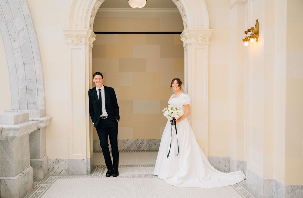 Bride in white gown holding bouquet and groom in black suit pose in elegant marble hall.