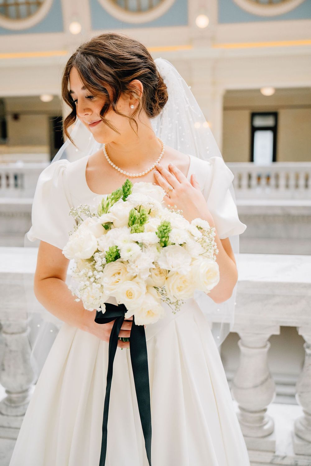 Bride in white gown with veil holds white floral bouquet with black ribbon at elegant marble venue.