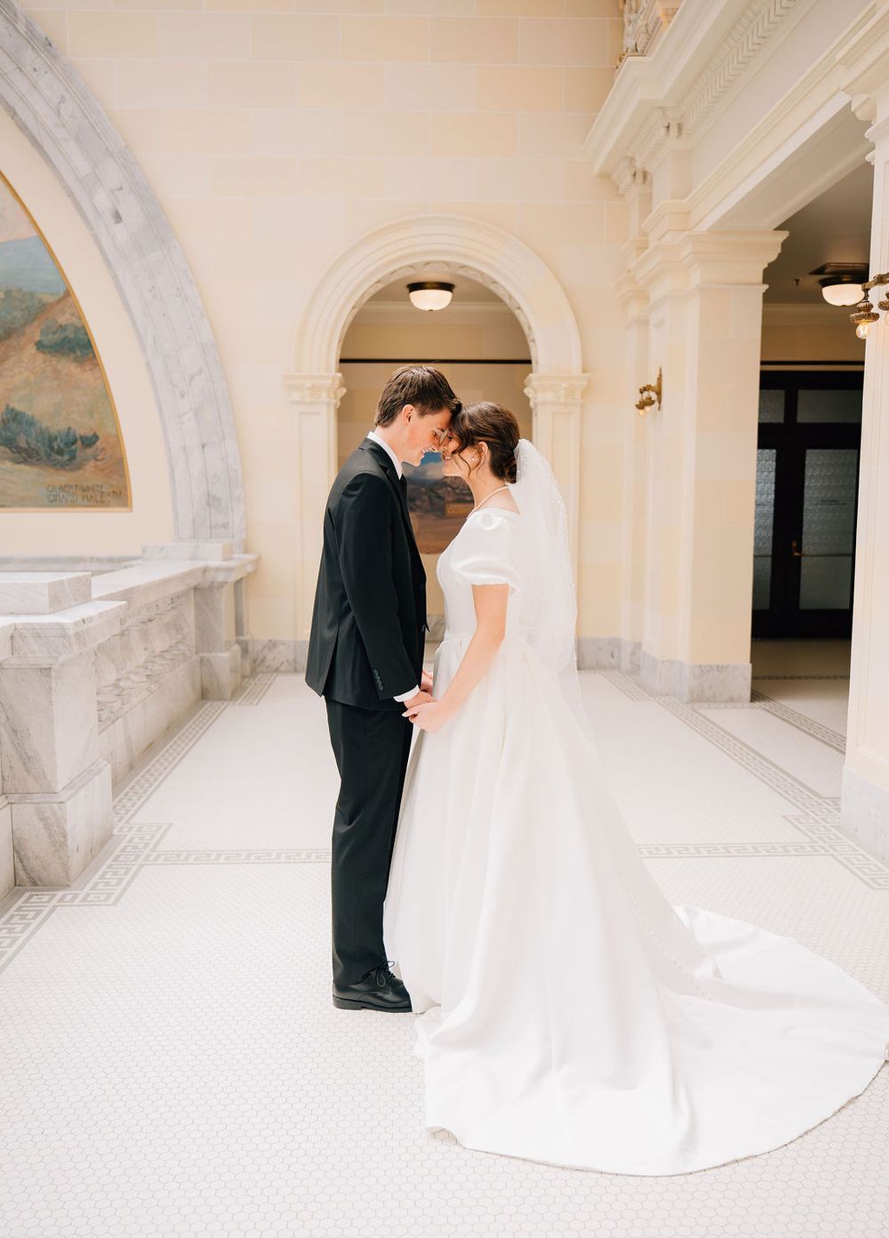 Bride and groom share a romantic forehead touch in an elegant arched hallway, bride in a flowing white gown with veil.