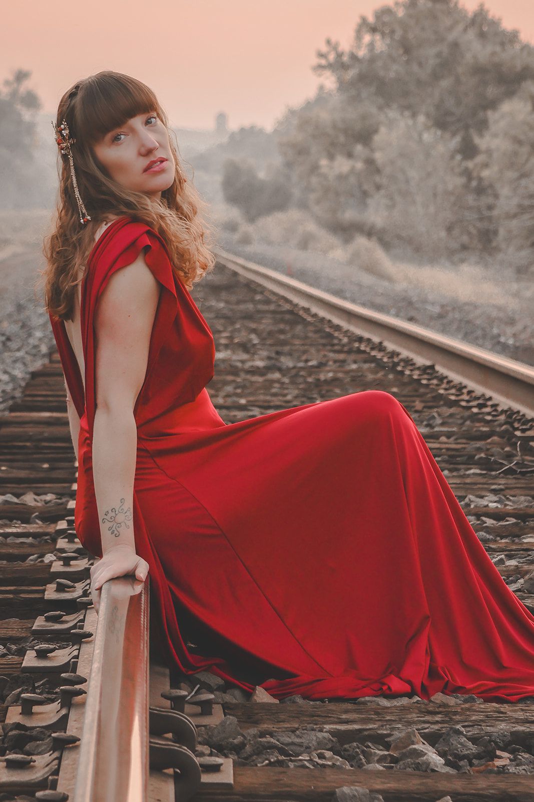 A person in a flowing red dress poses dramatically on train tracks during sunset, creating a moody atmosphere.