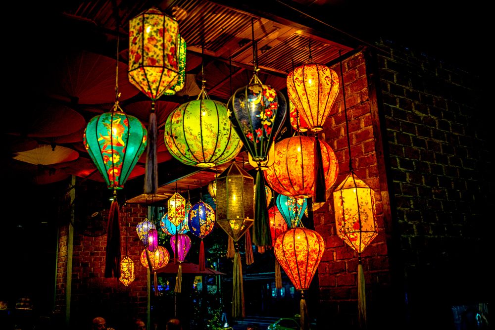 Colorful traditional Asian lanterns glowing in vibrant hues of orange, green, and red illuminate a dark space.